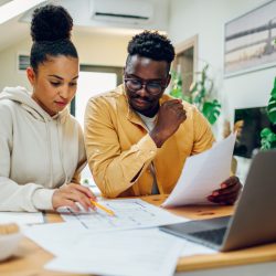 A couple sit at their kitchen table, paper spread before them. One points at a paper while the other views a laptop screen.