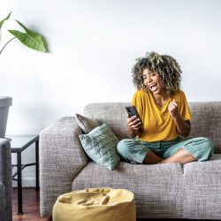 A woman sits on her couch, smiling and cheering as she looks down at her cell phone.