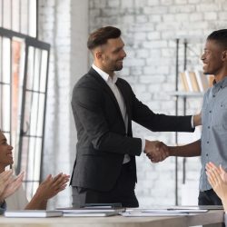 A boss and team member stand at the head of a conference table. The boss shakes the team member’s hand, while coworkers sitting at the table applaud the team member.