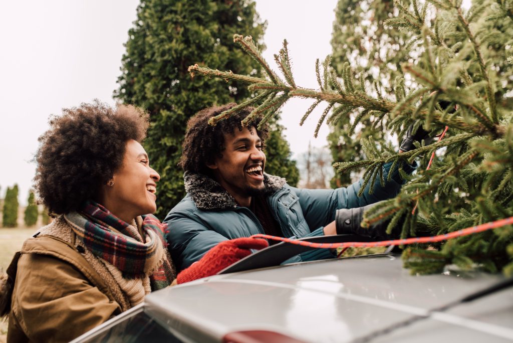 Couple ties a Christmas tree to the roof of their car. They smile at each other as they secure the tree.