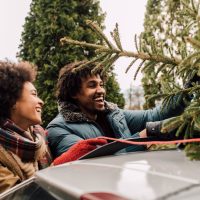 Couple ties a Christmas tree to the roof of their car. They smile at each other as they secure the tree.