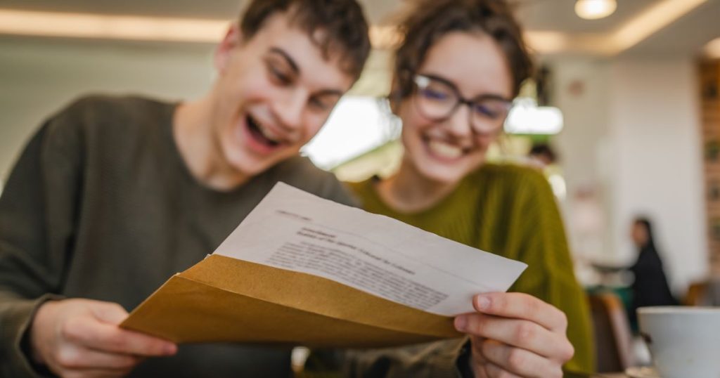 A teenage child pulls a college acceptance letter out of a large yellow envelope. A family member sits behind them, smiling in anticipation. 