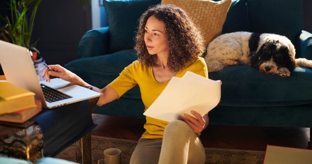 A person sits on the floor with their laptop building a budget, as their dog sleeps on the couch behind them.
