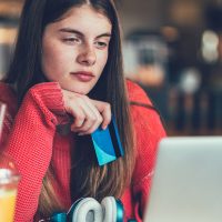 A college student sits in a coffee shop making online purchases with their laptop and credit card.
