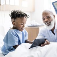 A grandfather shows a digital tablet to his sick grandson in a hospital bed. A nurse stands in the foreground taking vitals. 