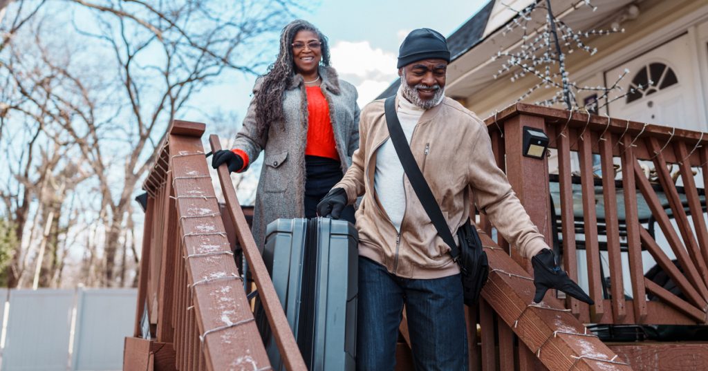 A couple walk down the porch stairs, suitcases in hand, leaving their house for a fun holiday getaway.