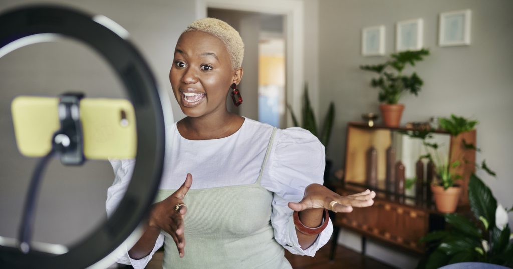 An influencer stands in front of a phone and ring light, recording a vlog from their home.