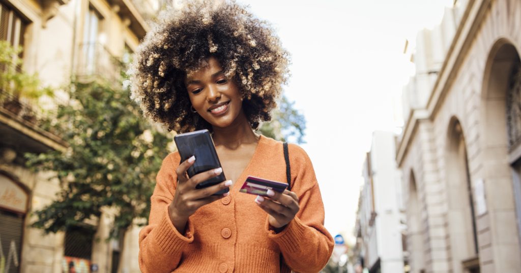 A woman stands in the street, holding her phone in one hand and her credit card in the other.