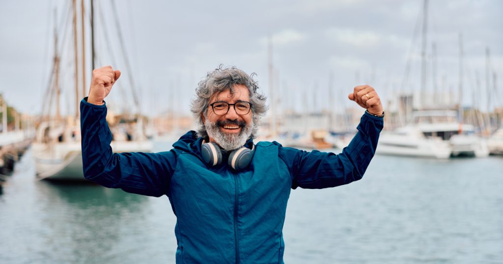Mature adult man wearing headphones around his neck, standing by a marina with boats, smiling at the camera and flexing his arms, showing strength.