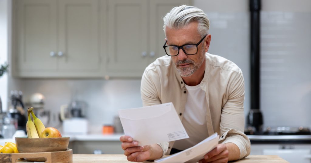 Man at home reading a bill he got in his mail at the kitchen counter.