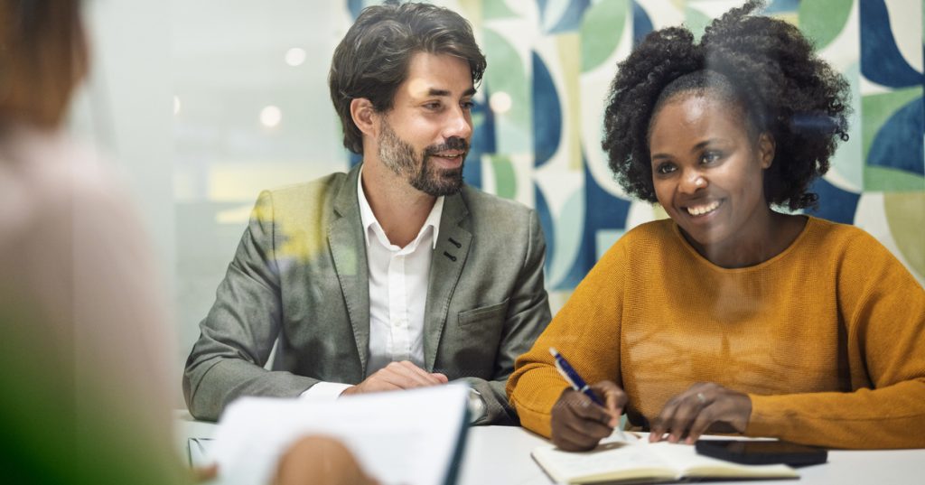 A couple sits next to each other at a table as they speak to a loan officer while applying for mortgage.