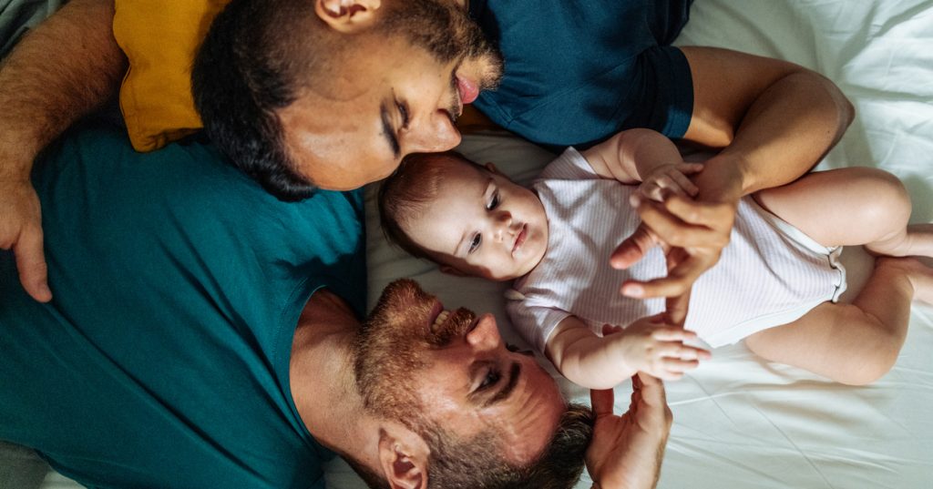 A couple lies in bed with their baby girl as they look at her smiling.