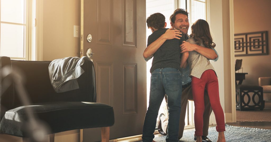 A father arrives home, bending down in the entryway to hug his son and daughter.