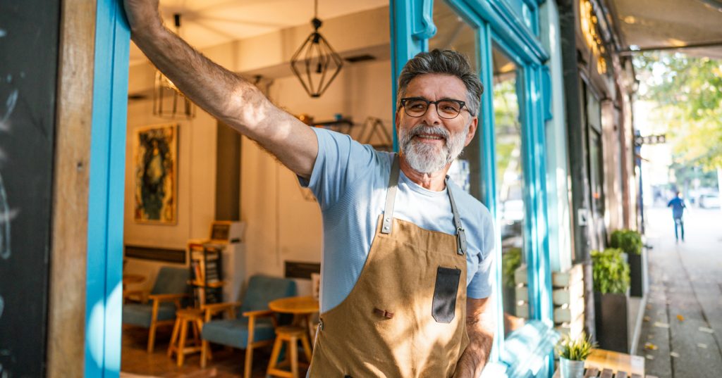 A coffee shop owner stands in the doorway of his business, leaning on the doorframe as he looks outside.