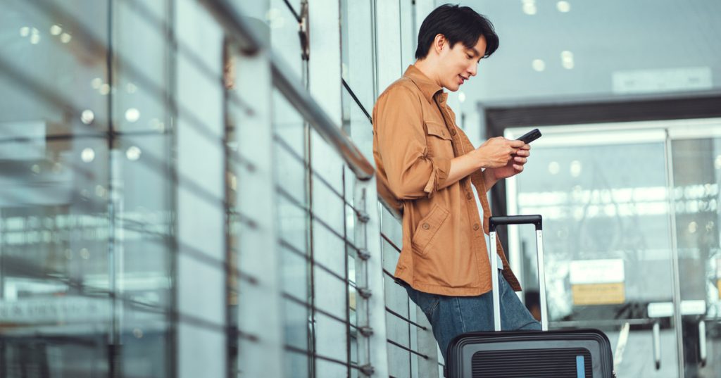 A man uses his mobile smartphone while standing with his suitcase inside an airport terminal.