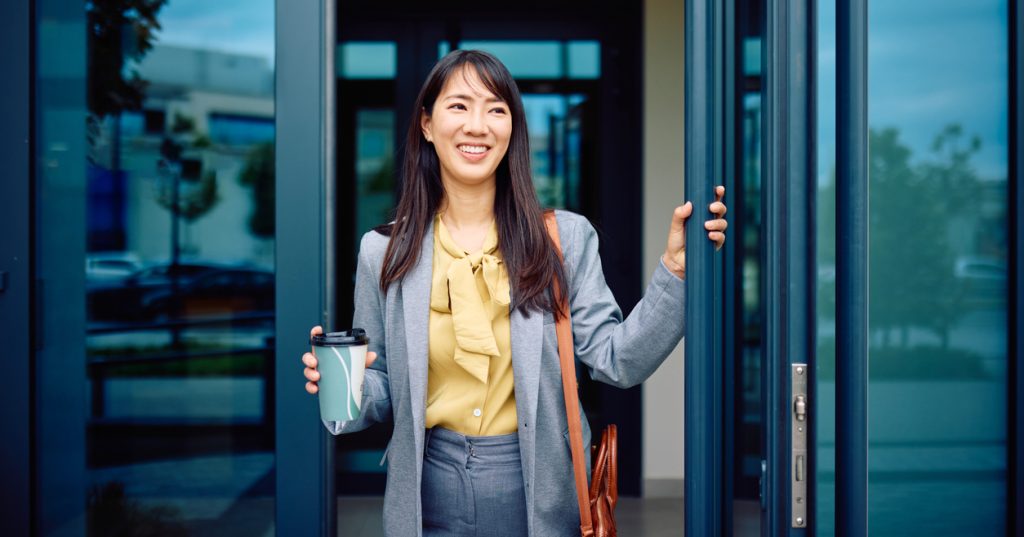 A young businesswoman is arriving at the office, opening the glass door while holding a coffee cup in her other hand.