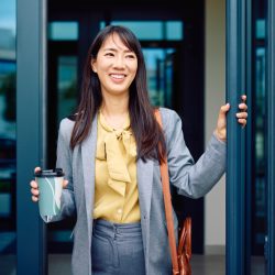 A young businesswoman is arriving at the office, opening the glass door while holding a coffee cup in her other hand.