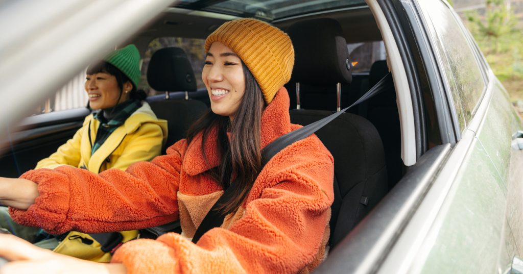 Photo of two friends driving down the road, chatting and enjoying their trip. They are both wearing bright, fall-colored jackets.