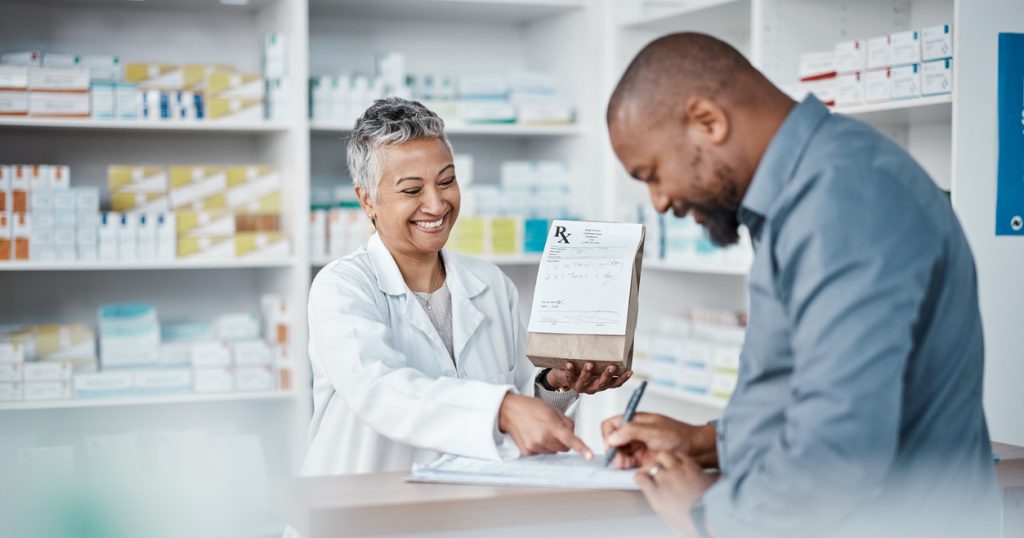 A pharmacist hands a bag of medicine to a man as he finishes filling out paperwork at the pharmacy counter.